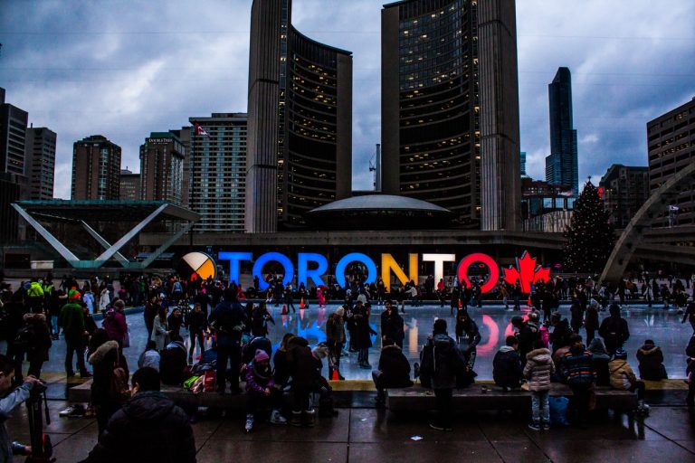 a night-time picture of downtown Toronto with buildings in the background and people near the fountain