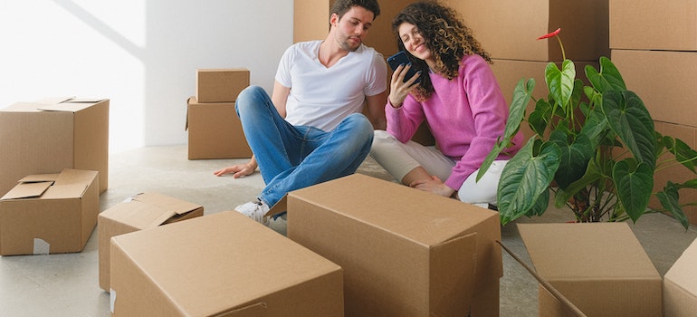 A couple surrounded by moving boxes looking at a phone and smiling