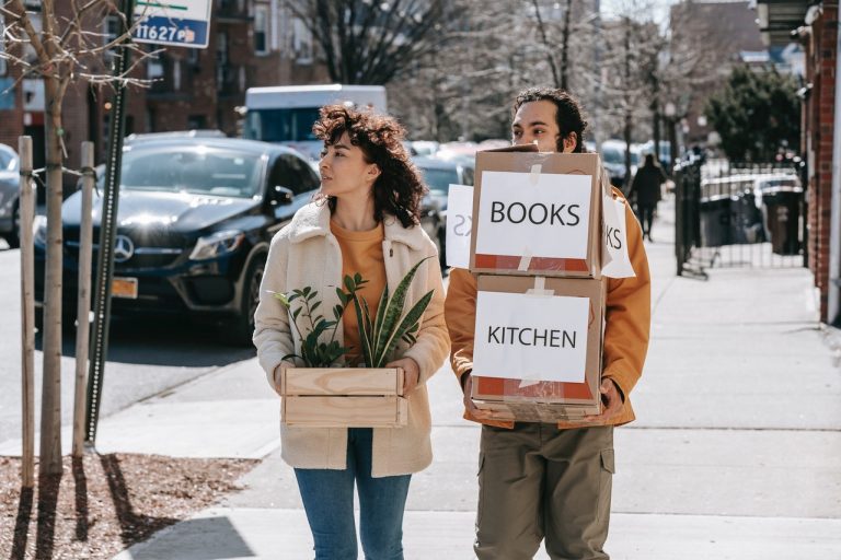 Couple walking on sidewalk carrying boxes and plants