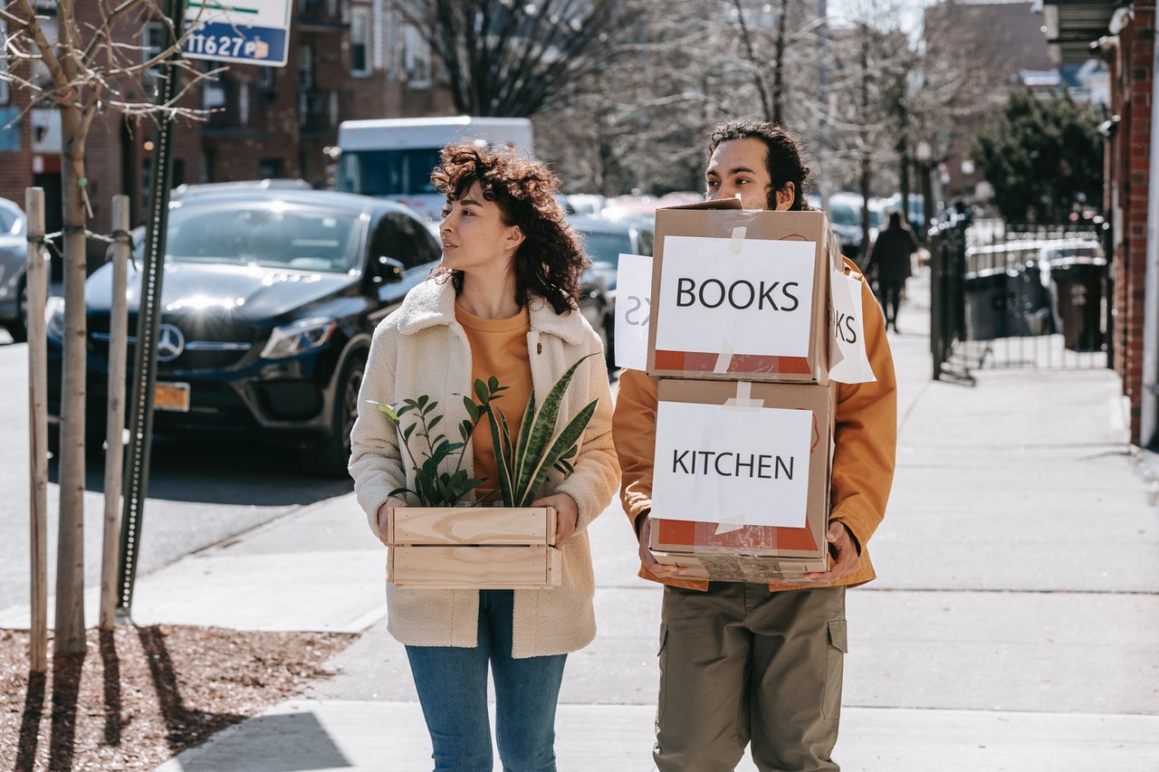 Couple walking on sidewalk carrying boxes and plants