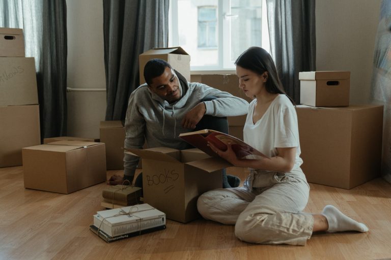 Men and woman in white clothes, surrounded with moving boxes are sitting on the floor and reading.
