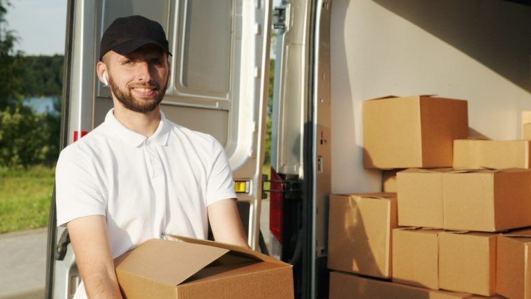 Man smiling while holding a box in front of a moving van full of boxes
